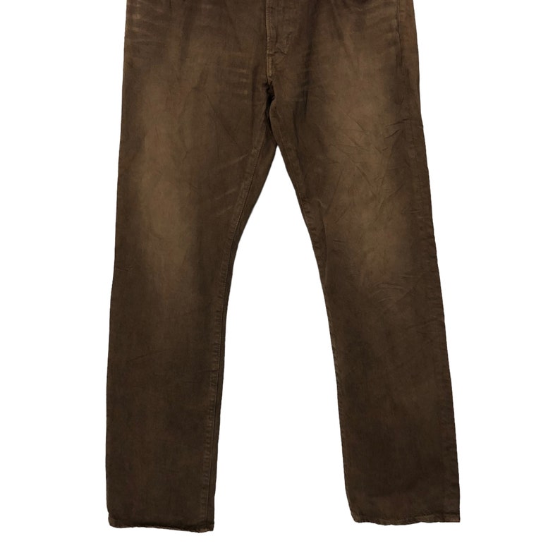May include: Brown denim jeans with a classic five-pocket design. The pants have a straight leg cut and a button and zipper closure. The fabric has a slightly faded appearance, giving them a worn-in look.