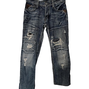 Op de afbeelding: Donkerblauwe spijkerbroek van denim met een verwassen look, voorzien van meerdere scheuren en gerafelde plekken op de voorzijde van de broekspijpen. De broek heeft een knoopsluiting, vijf zakken met bruine lederachtige accenten en stiksels.