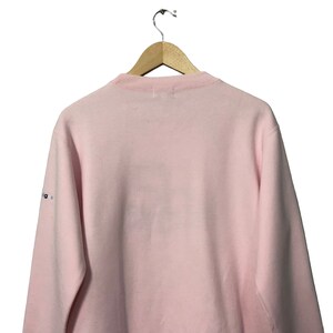 May include: A light pink sweatshirt with a crew neck, hanging from a wooden hanger. The back of the sweatshirt is visible, with a small logo on the left sleeve. The fabric appears soft and comfortable.