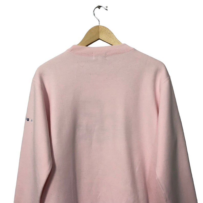 May include: A light pink sweatshirt with a crew neck, hanging from a wooden hanger. The back of the sweatshirt is visible, with a small logo on the left sleeve. The fabric appears soft and comfortable.