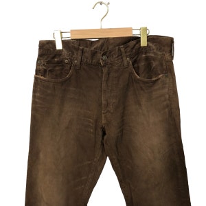 May include: Brown corduroy pants hanging on a wooden hanger. The pants have a classic five-pocket design and a button closure. The fabric has a textured appearance with subtle variations in color.