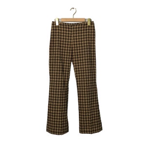Può includere: Pantaloni con motivo a pied-de-poule marrone e nero appesi a una gruccia di legno. I pantaloni hanno un design a gamba svasata e sono realizzati in un tessuto tessuto. Il motivo è un design classico e ripetitivo.