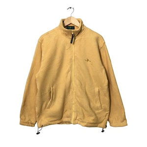 May include: A yellow zip-up fleece jacket with a high collar and two front pockets. The jacket has a textured waffle-like pattern and a small embroidered logo on the chest. The jacket is hanging on a wooden hanger.