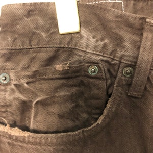 May include: Close-up of brown denim jeans, showing the front pocket and metal rivets. The jeans have a worn appearance with visible distressing around the pocket edges. The rivets have a circular design.