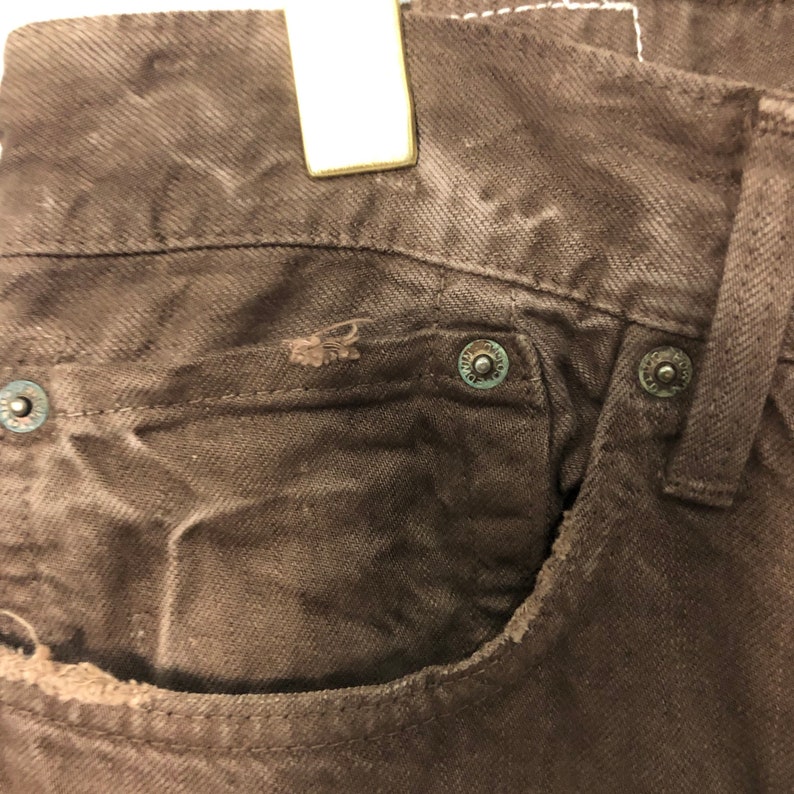 May include: Close-up of brown denim jeans, showing the front pocket and metal rivets. The jeans have a worn appearance with visible distressing around the pocket edges. The rivets have a circular design.