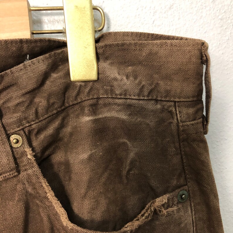 May include: Close-up of brown denim pants with a distressed pocket edge. The pants are hanging from a gold-colored hanger. The fabric shows signs of wear and fading, with metal rivets and a button visible.