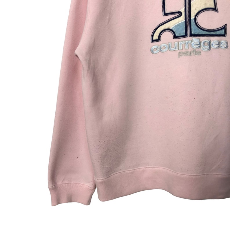 May include: A light pink sweatshirt with the text "courreges paris" in silver and a logo in blue, white, and black. The sweatshirt has a crew neck and long sleeves.