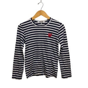 Puede incluir: Una camiseta de manga larga a rayas con rayas horizontales azul marino y blancas. La camiseta presenta un pequeño logotipo de corazón rojo con ojos en el pecho izquierdo. La camiseta está colgada de una percha de madera.