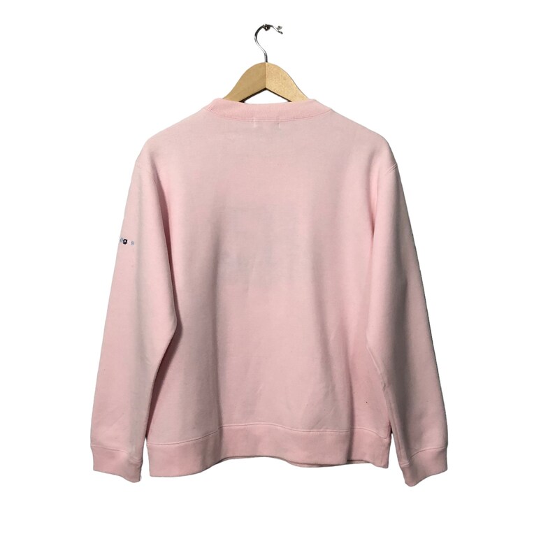 May include: A light pink sweatshirt with a crew neck, hanging on a wooden hanger. The long-sleeved top has a simple design, with a small logo on the left sleeve. The sweatshirt is made of a soft material.