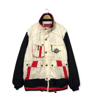 May include: A multi-pocket jacket featuring a cream-colored front panel with red and black accents, black sleeves, and a high collar. The front includes a compass graphic, multiple snap-button pockets, and a drawstring hem with red cord toggles.