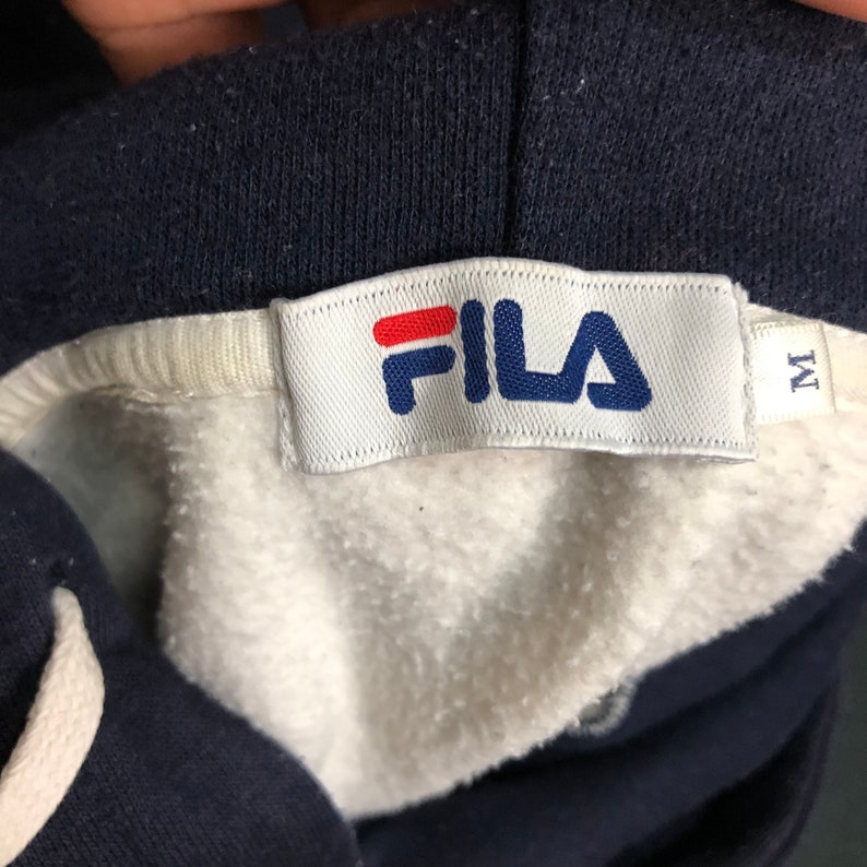 May include: A navy blue hooded sweatshirt with a white interior lining and a white fabric tag featuring the red and blue Fila logo. A smaller white tag attached to the side indicates a size M. The garment includes a white drawstring cord.