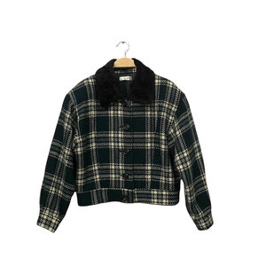 Peut inclure: Veste à carreaux verts et blancs avec un col en fausse fourrure noire. La veste a une fermeture boutonnée et des manches longues. Elle est suspendue à un cintre en bois.