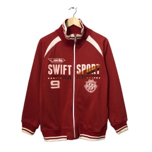 May include: A maroon zip-up track jacket with white and cream accents. The jacket features the words "SWIFT SPORT" in large white letters, along with the number 9 and other graphics. The jacket has a ribbed collar, cuffs, and hem.