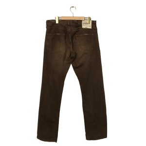 May include: Brown denim jeans hanging on a wooden hanger. The jeans have a classic five-pocket design with a small red tag on the back pocket. The brand name "EDWIN" is visible on a tag.