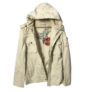 Könnte beinhalten: Cremefarbene Kapuzenjacke mit mehreren Taschen und einem Reißverschluss vorne. Die Jacke hat eine Grafik auf der Innenseite mit einem Adler und dem Text "SKIN WHOOP LIKE AN EAGLE". Die Jacke hängt an einem Holzkleiderbügel.