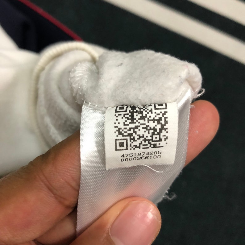 May include: A white fabric clothing tag featuring a black and white QR code and the numerical sequence 4751874205 0000366100 printed in black ink. The tag is attached to a white fleece garment with a soft, textured interior lining.