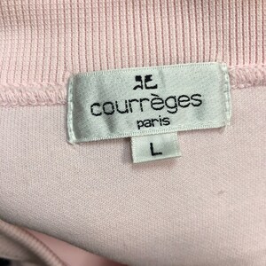 May include: Close-up of a light pink garment tag with the brand name "courr&egrave;ges paris" in black text. The tag also features a stylized logo and the size "L". The garment appears to be a light pink knit fabric.