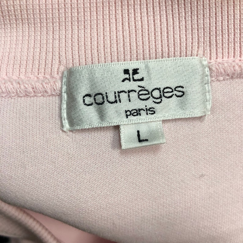 May include: Close-up of a light pink garment tag with the brand name "courr&egrave;ges paris" in black text. The tag also features a stylized logo and the size "L". The garment appears to be a light pink knit fabric.