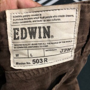 May include: Close-up of a brown fabric with a rectangular label. The label reads "EDWIN" in bold letters, with text about the brand's mission. The label also includes "Mission No. 503 R" and "JPN" for nationality.