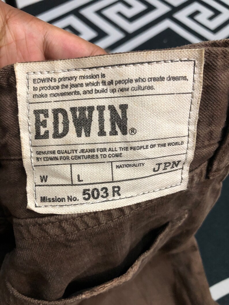 May include: Close-up of a brown fabric with a rectangular label. The label reads "EDWIN" in bold letters, with text about the brand's mission. The label also includes "Mission No. 503 R" and "JPN" for nationality.