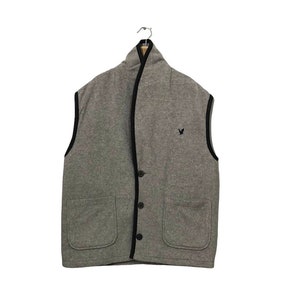 May include: A sleeveless grey vest featuring a shawl collar, a front button closure with three dark buttons, and two large patch pockets. The garment is finished with black trim along the edges and displays a small black eagle logo on the chest.
