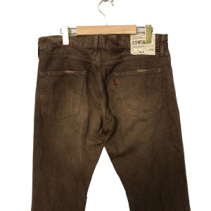 May include: Brown denim jeans with a distressed look, featuring back pockets and a red logo tag. The jeans are hanging on a wooden hanger. The brand tag reads "EDWIN".