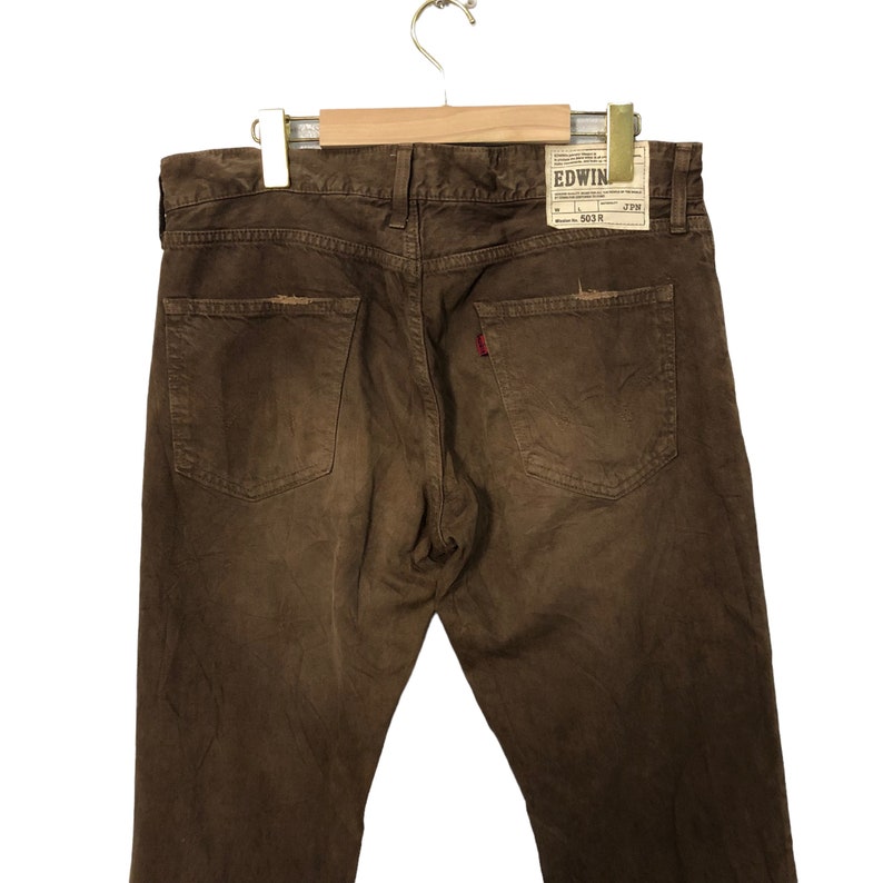 May include: Brown denim jeans with a distressed look, featuring back pockets and a red logo tag. The jeans are hanging on a wooden hanger. The brand tag reads "EDWIN".