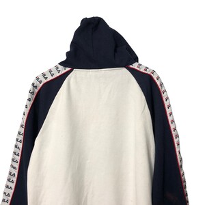 May include: A navy blue and white hooded sweatshirt featuring raglan sleeves with a repeating FILA logo tape running down the length of each arm. The hoodie has a solid navy blue hood and dark blue sleeves, contrasted by a white central body panel.