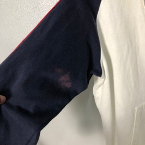 May include: A long-sleeve sweatshirt featuring a white torso and dark navy blue sleeves. A thin red stripe runs along the top of the navy sleeves. There is a noticeable reddish-brown stain on the upper portion of the dark navy blue sleeve fabric.