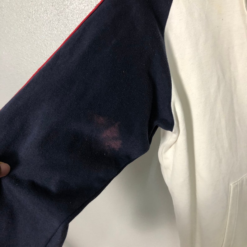 May include: A long-sleeve sweatshirt featuring a white torso and dark navy blue sleeves. A thin red stripe runs along the top of the navy sleeves. There is a noticeable reddish-brown stain on the upper portion of the dark navy blue sleeve fabric.