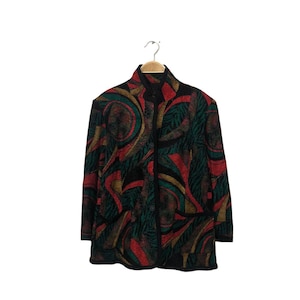 May include: A button-down jacket with a vibrant abstract pattern in red, green, and gold on a black background. The jacket is hanging on a wooden hanger against a white backdrop. The jacket has a high collar and two pockets.