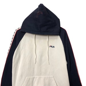 May include: A navy blue and off-white hooded sweatshirt featuring a raglan sleeve design. The front displays the Fila logo in red and blue embroidery. The sleeves are accented with a white side stripe containing small navy blue triangular patterns.