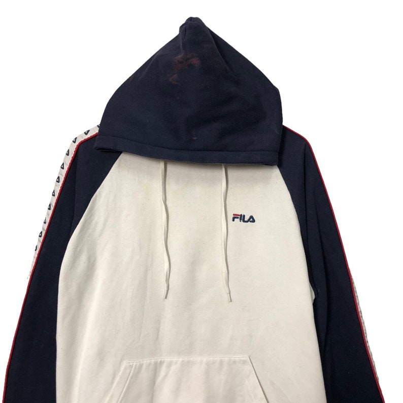 May include: A navy blue and off-white hooded sweatshirt featuring a raglan sleeve design. The front displays the Fila logo in red and blue embroidery. The sleeves are accented with a white side stripe containing small navy blue triangular patterns.
