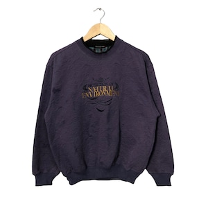 Puede incluir: Sudadera morada oscura con el texto dorado "PROTECTION OF NATURAL ENVIRONMENT". La sudadera tiene un patrón texturizado y cuello y puños acanalados azul oscuro. Cuelga de una percha de madera.