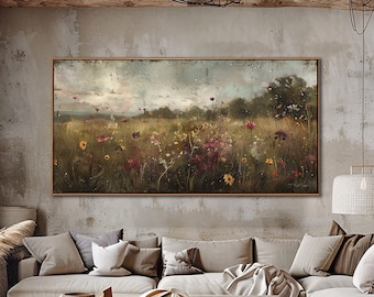May include: A large rectangular framed painting shows a field of wildflowers in yellow, red, and purple hues. The artwork hangs above a light-coloured sectional sofa with several cushions. The room has a rustic, natural aesthetic.