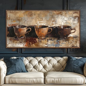 May include: An abstract painting of three brown coffee cups with handles, set against a textured background of beige, brown, and white. The artwork hangs above a cream-colored tufted sofa with blue pillows, a wooden side table, and a lamp.