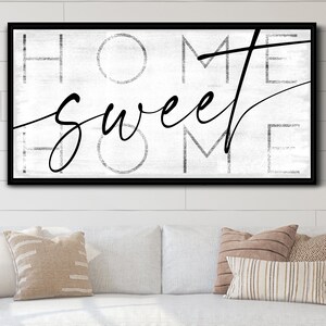 May include: A framed wall art piece with the words "HOME sweet HOME" in black script and block letters. The background is a distressed white, and the frame is black. The artwork is displayed above a white sofa with decorative pillows.