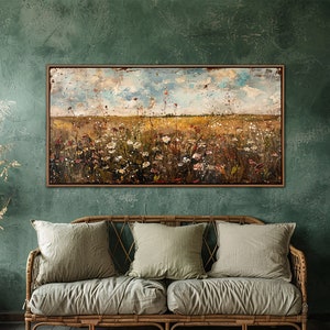 Puede incluir: Una pintura abstracta de paisaje enmarcada representa un campo de flores silvestres bajo un cielo nublado. Cuelga sobre un sof&aacute; de mimbre con tres cojines verde salvia, creando una est&eacute;tica r&uacute;stica y natural.