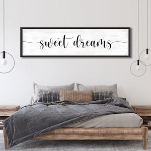 Op de afbeelding: Een zwart ingelijst bord met een witte achtergrond en de tekst "sweet dreams" in zwarte cursieve letters. Het bord hangt boven een bed met een grijs dekbed en een bruin houten frame. Twee zwarte hanglampen met ronde lampenkappen hangen aan het plafond.