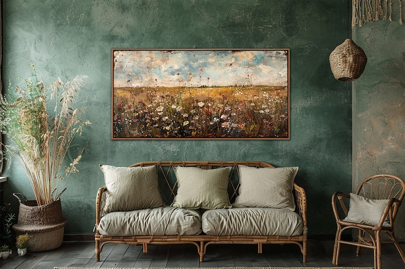 Puede incluir: Una pintura abstracta de paisaje enmarcada representa un campo de flores silvestres bajo un cielo nublado. Cuelga sobre un sof&aacute; de mimbre con tres cojines verde salvia, creando una est&eacute;tica r&uacute;stica y natural.