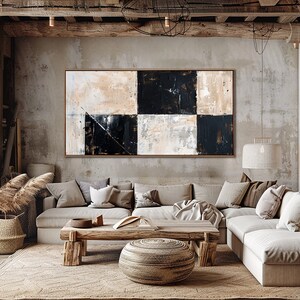 May include: A large abstract painting with black and white squares on a beige background hangs above a sectional sofa in a living room. The sofa is covered in white fabric and has several throw pillows. The room is decorated in a rustic style with exposed wooden beams and a concrete floor.