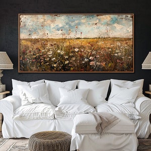 Puede incluir: Una pintura de paisaje abstracto en un marco de madera, que representa un campo de flores silvestres bajo un cielo nublado. La pintura cuelga sobre un sof&aacute; blanco con m&uacute;ltiples almohadas. Dos l&aacute;mparas con pantallas beige flanquean la pintura.