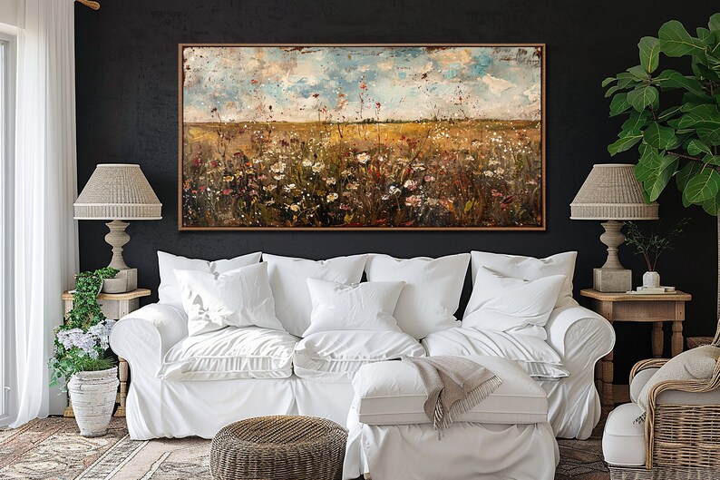 Puede incluir: Una pintura de paisaje abstracto en un marco de madera, que representa un campo de flores silvestres bajo un cielo nublado. La pintura cuelga sobre un sof&aacute; blanco con m&uacute;ltiples almohadas. Dos l&aacute;mparas con pantallas beige flanquean la pintura.