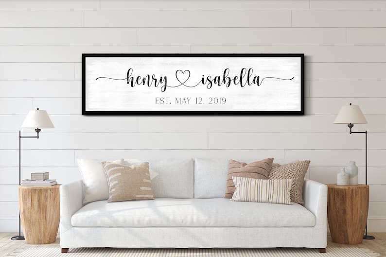 May include: A black framed sign with a white background and black text. The text reads "henry &hearts; isabella EST. MAY 12, 2019". The sign is hanging on a white wall above a white sofa with brown throw cushions.