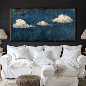 May include: A white couch with white pillows and a throw blanket sits in front of a dark blue abstract painting with white clouds. The painting is framed in wood. The couch is in a living room with a side table and a lamp.