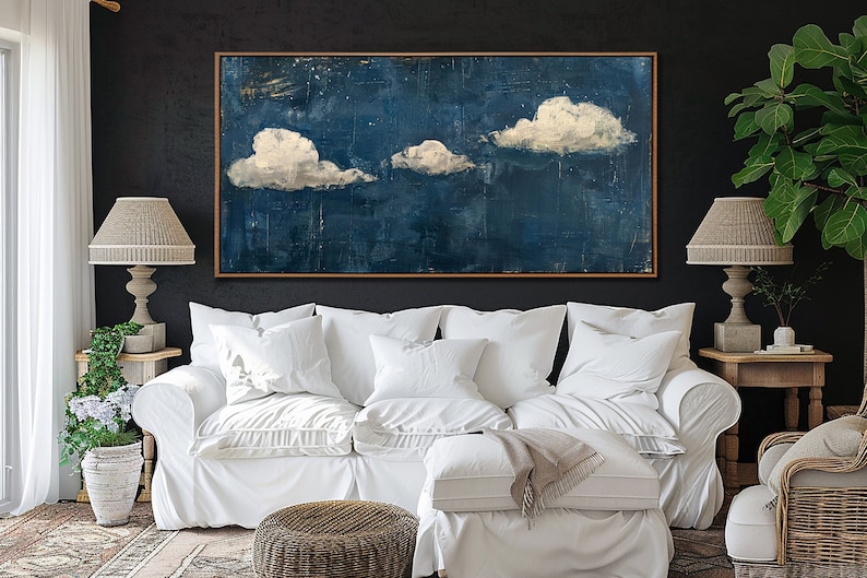 May include: A white couch with white pillows and a throw blanket sits in front of a dark blue abstract painting with white clouds. The painting is framed in wood. The couch is in a living room with a side table and a lamp.