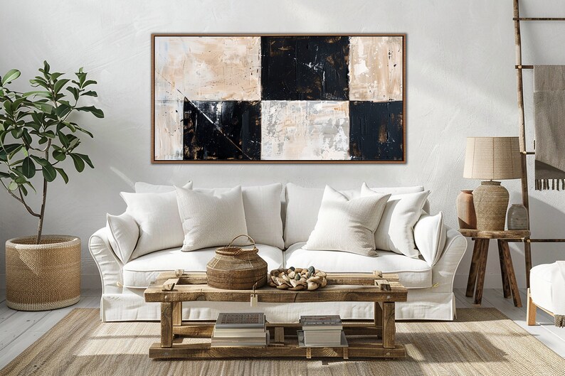 May include: A white sofa with two pillows sits in front of a large abstract painting with black and white squares. The painting is framed in a light brown wood frame. A wooden coffee table with a woven basket and a bowl of stones sits in front of the sofa.