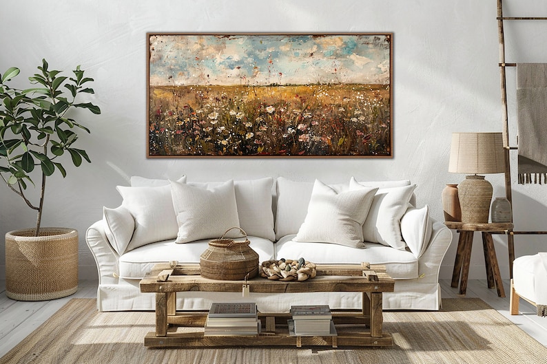 Puede incluir: Una pintura de paisaje abstracto en un marco de madera. La obra de arte representa un campo de flores silvestres bajo un cielo nublado. La paleta de colores incluye tonos de marr&oacute;n, amarillo, blanco y azul. La pintura se exhibe sobre un sof&aacute; blanco.
