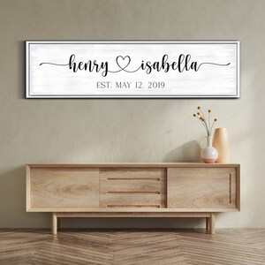 May include: A white wooden sign with black text that reads "henry & isabella est. may 12, 2019". The sign is hanging on a wall above a wooden dresser.