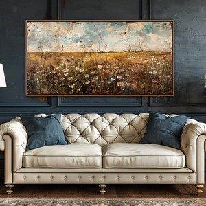 Puede incluir: Una pintura al &oacute;leo de un campo de flores silvestres en un marco marr&oacute;n cuelga sobre un sof&aacute; crema con dos cojines azules. Una l&aacute;mpara dorada y un jarr&oacute;n de flores se encuentran a ambos lados del sof&aacute;. La pared es azul oscuro.